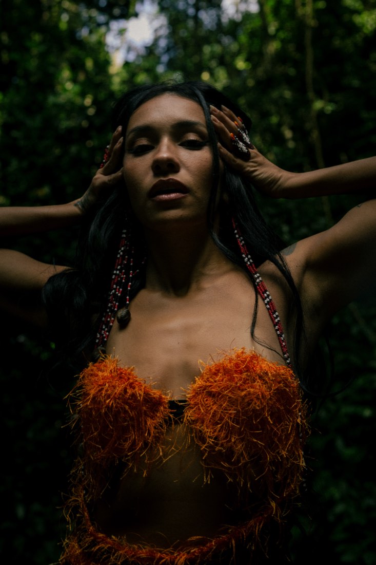 Marcele de corpo inteiro na mata, com flores laranja no peito.