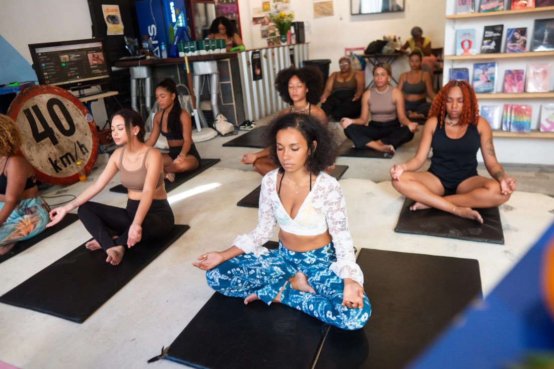 Mulheres sentadas em meditação sobre tapetes pretos num espaço com livros e uma placa de 40 km/h, no encontro do Projeto SerMente.
