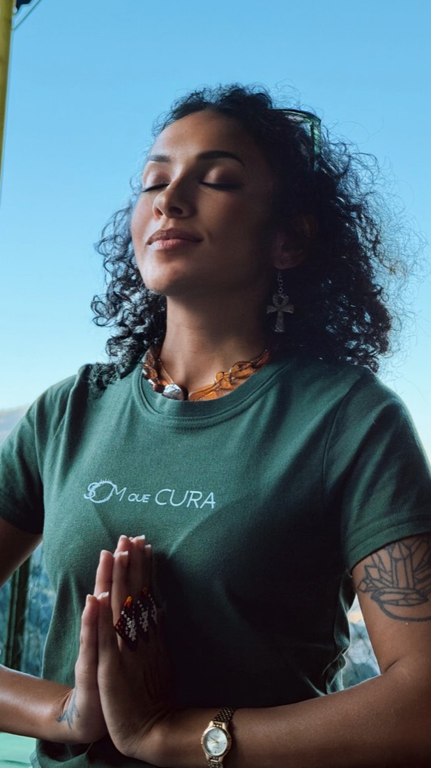 Marcele de olhos fechados e mãos em prece, vestindo a camiseta verde do Som que Cura, com o céu azul ao fundo.